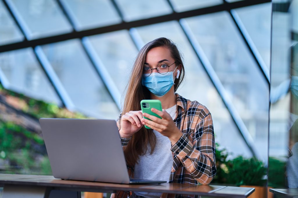 Girl wearing a mask, holding a phone, sitting and working with a notebook.