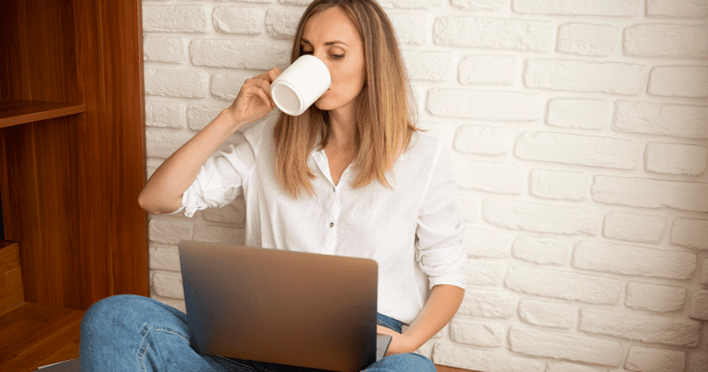 Young woman sitting on the floor, drinking a coffee and working on a laptop, with a relaxed and focused expression.