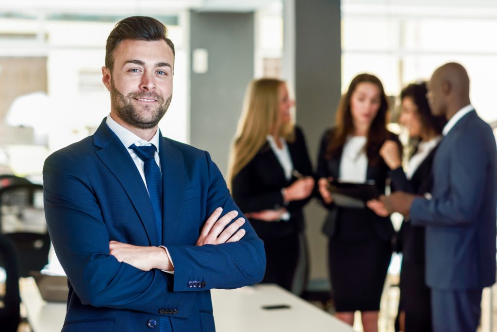 Smiling businessman in a suit with arms crossed, standing in front of a group having a discussion.