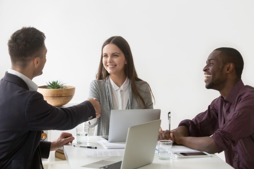 Smiling businesswoman with two male colleagues, one shaking hands and the other laughing, in an office setting.